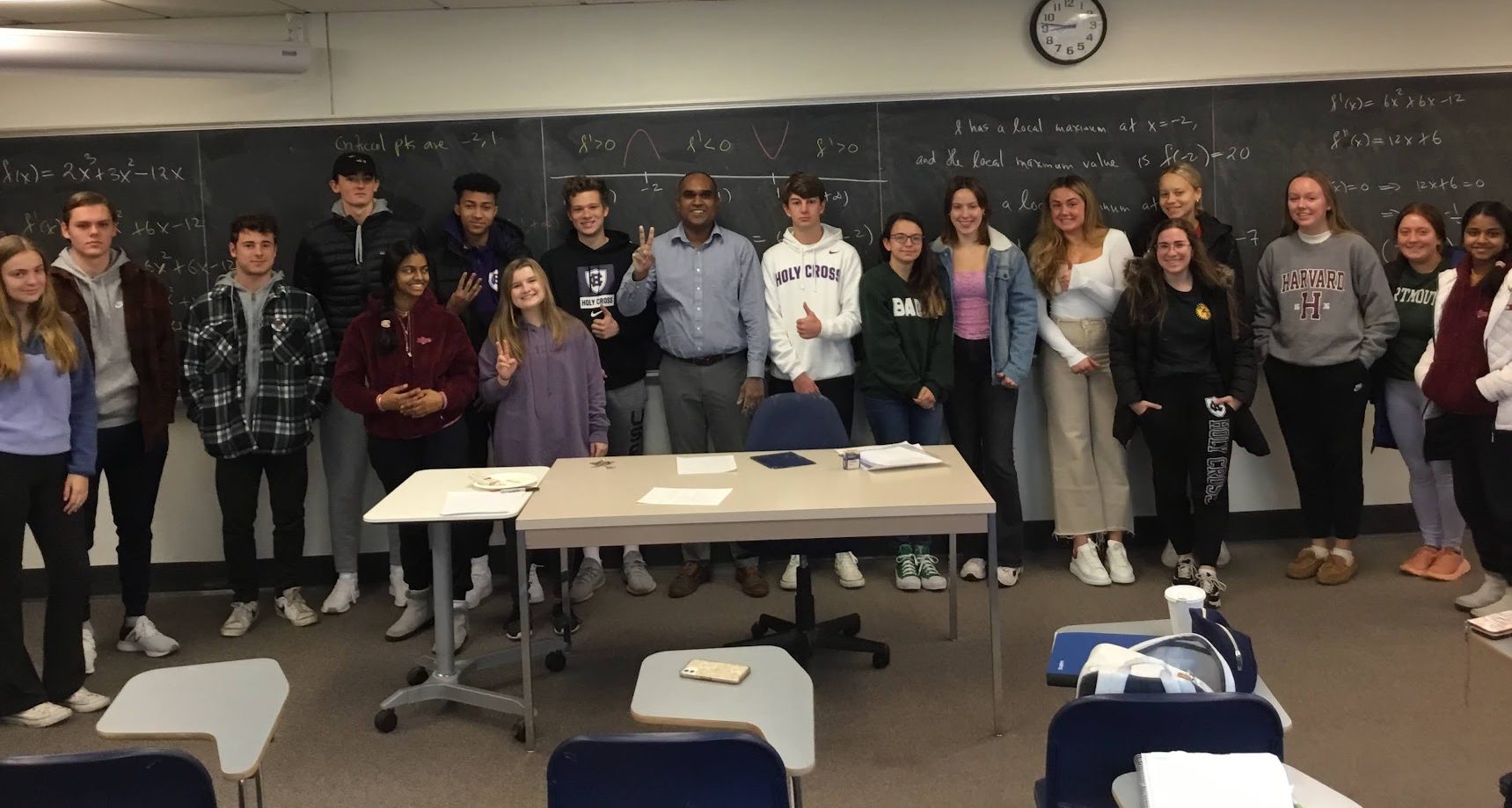 a group of students with their professor stands in front of a black chalkboard, covered in mathematical terms. All of the students are smiling, and some are holding up peace signs.